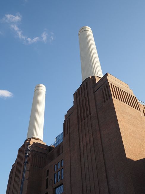 A low-angle view of the facade of Battersea Power Station, showcasing its red brick exterior with numerous rectangular windows, some with metal grids. Two large, cylindrical chimneys extend vertically